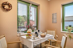 Un coin repas avec une table dressée pour deux personnes, une fenêtre donnant sur un immeuble et un panneau avec du texte accroché au mur.