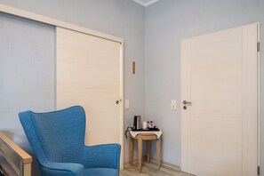 Una habitació amb una cadira blava, dues portes blanques, una taula petita amb una cafetera i un interruptor de llum fixat a la paret.