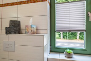A bathroom with a tiled wall, a wicker basket, a tissue box, a potted plant, and a window with blinds.