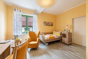 Une chambre d’hôtel de taille réduite, comprenant un lit, un bureau, une chaise, une table de chevet, une lampe et une fenêtre avec des rideaux.