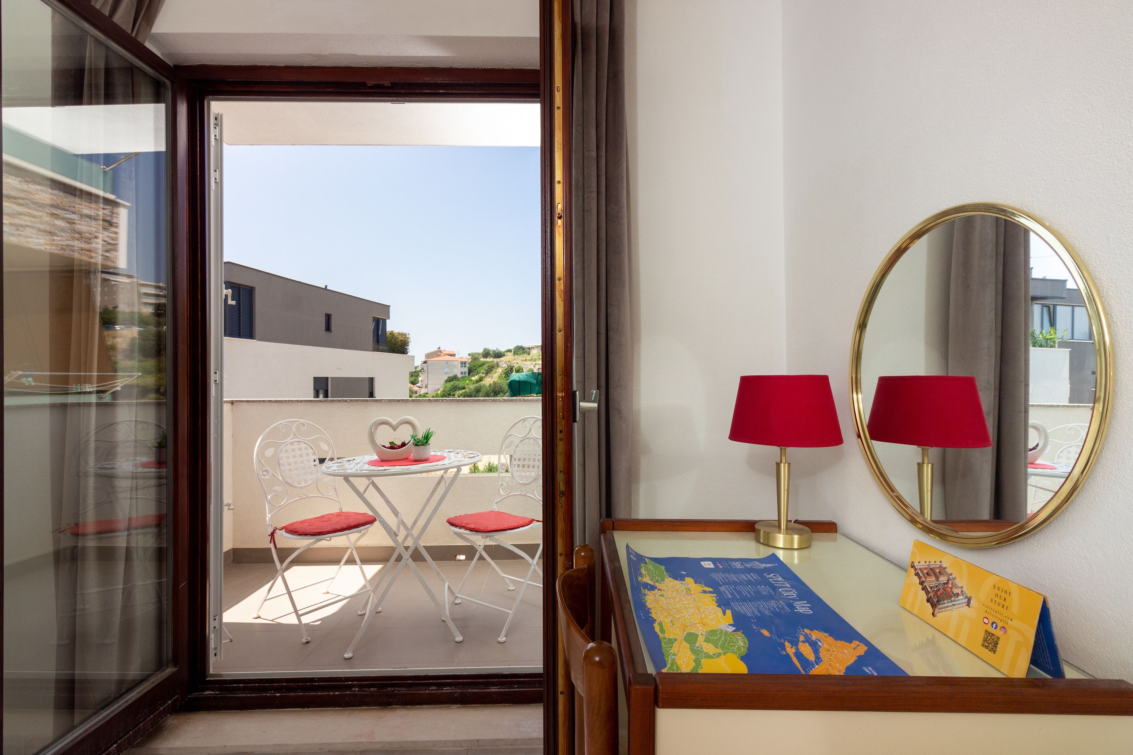 A room with a balcony, a table with chairs, a red lamp, a mirror, and a map on the wall.