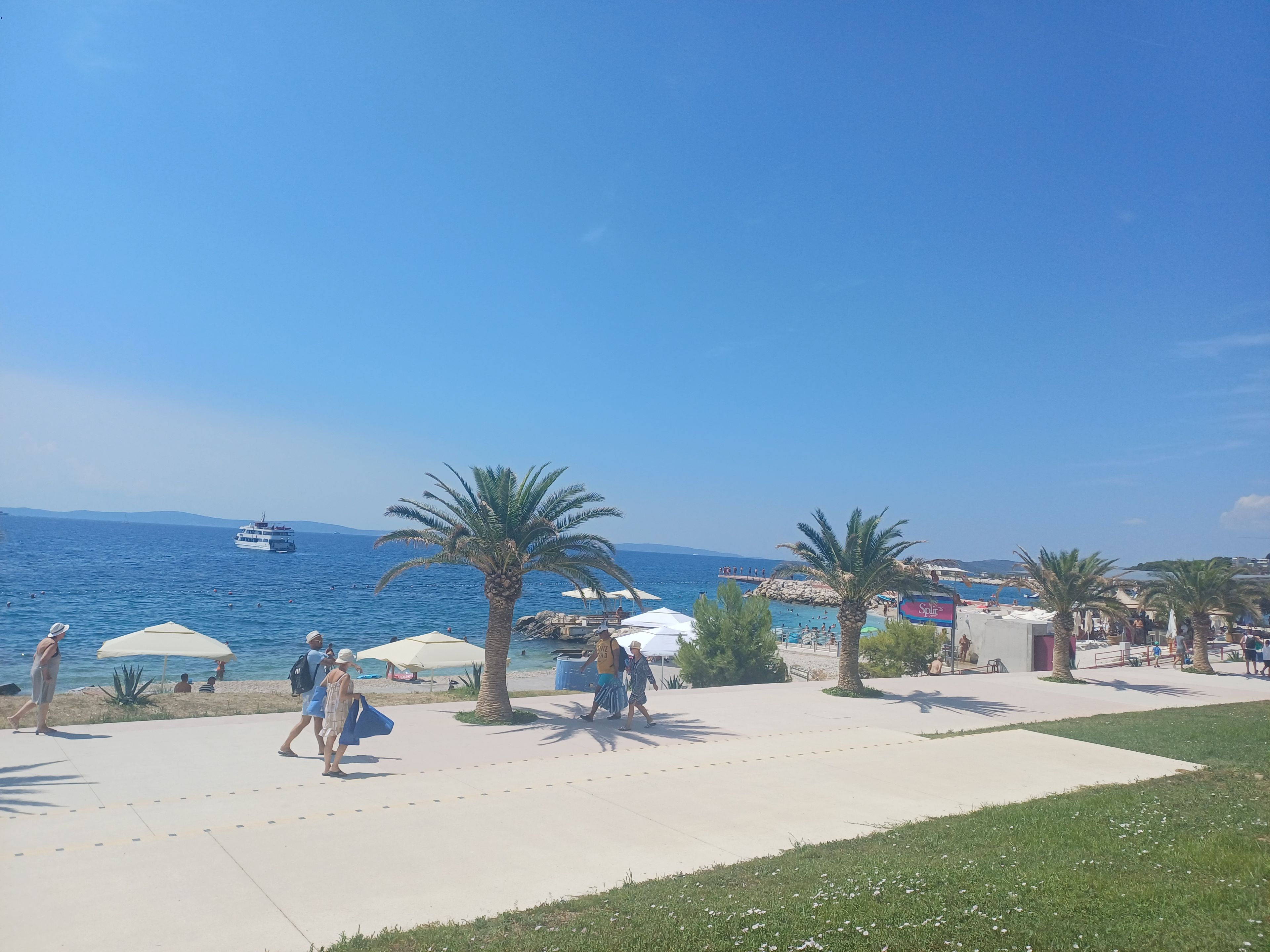 A coastal promenade with palm trees, umbrellas, and a clear blue sky.