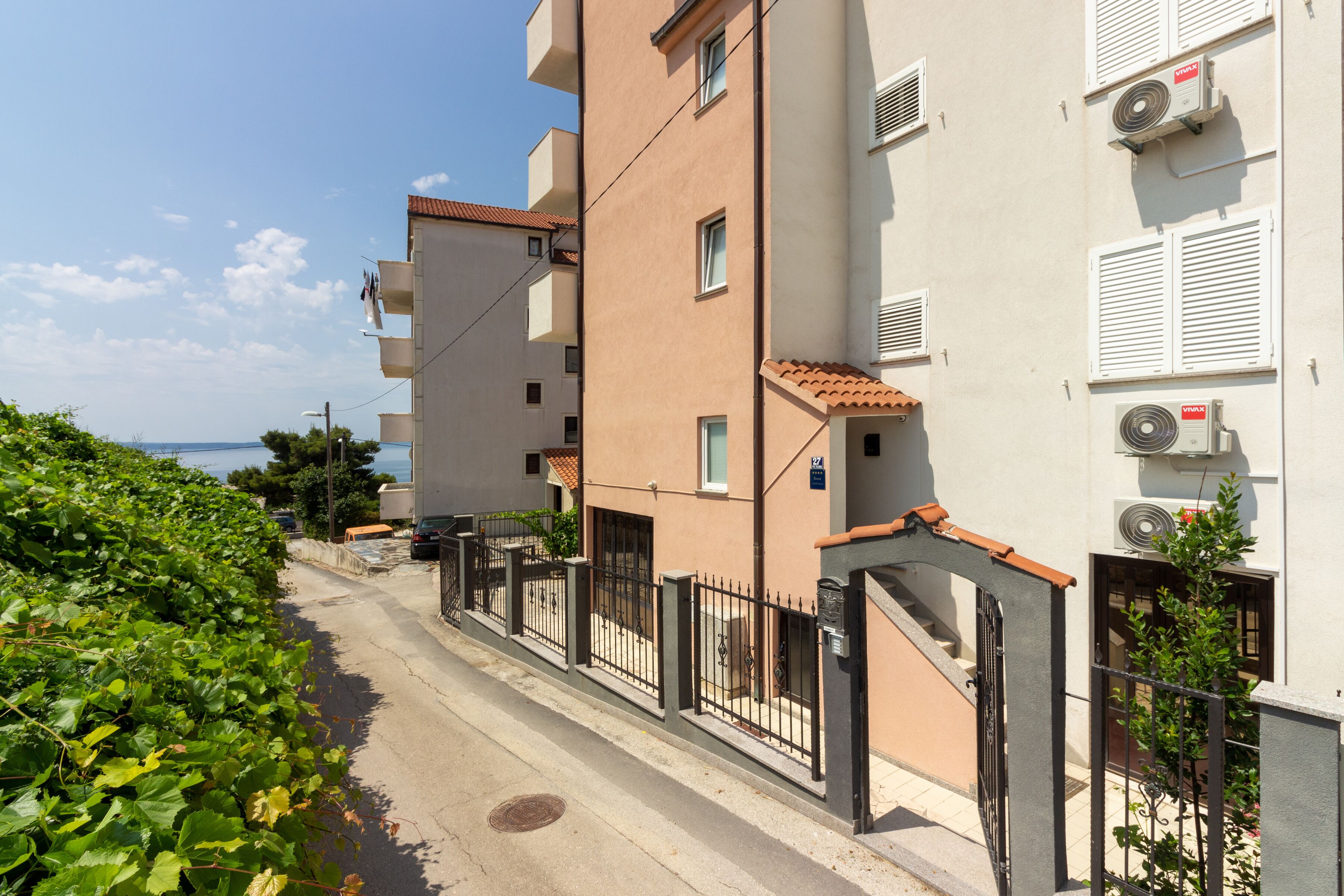 A narrow street with a building featuring balconies, air conditioning units, and a gate.