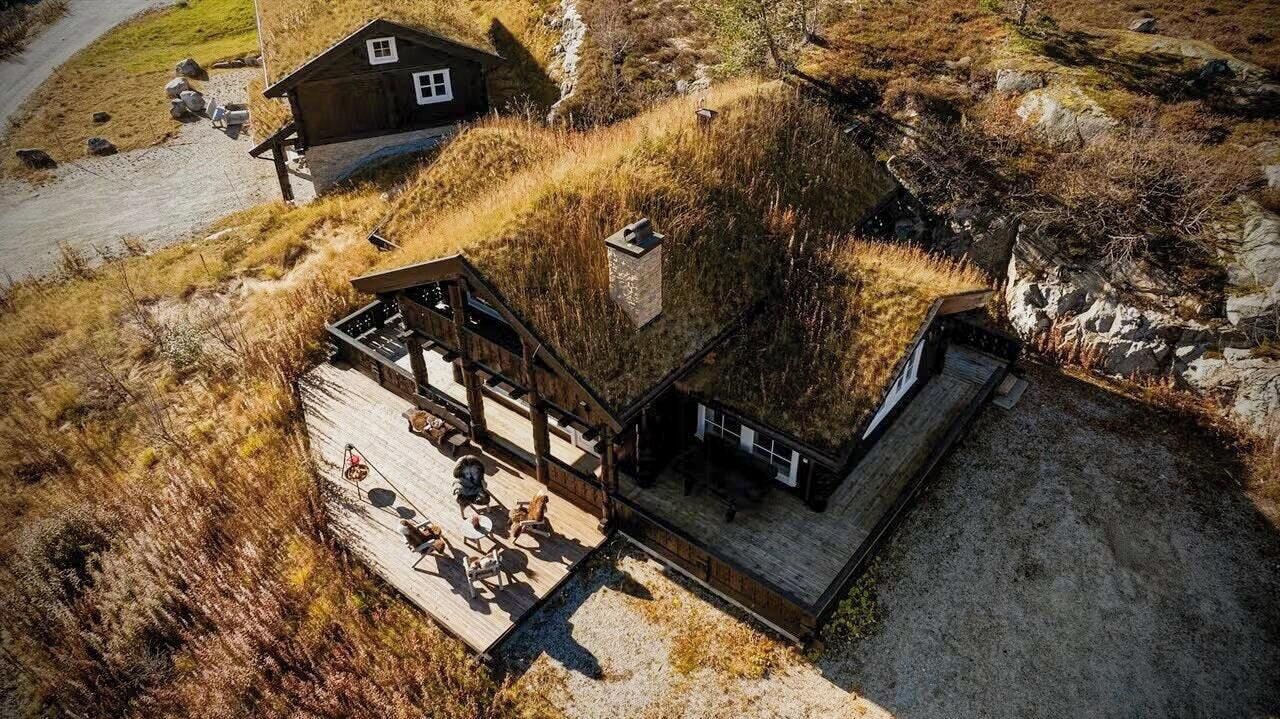 A wooden cabin with a deck and a small attached building, surrounded by natural terrain.