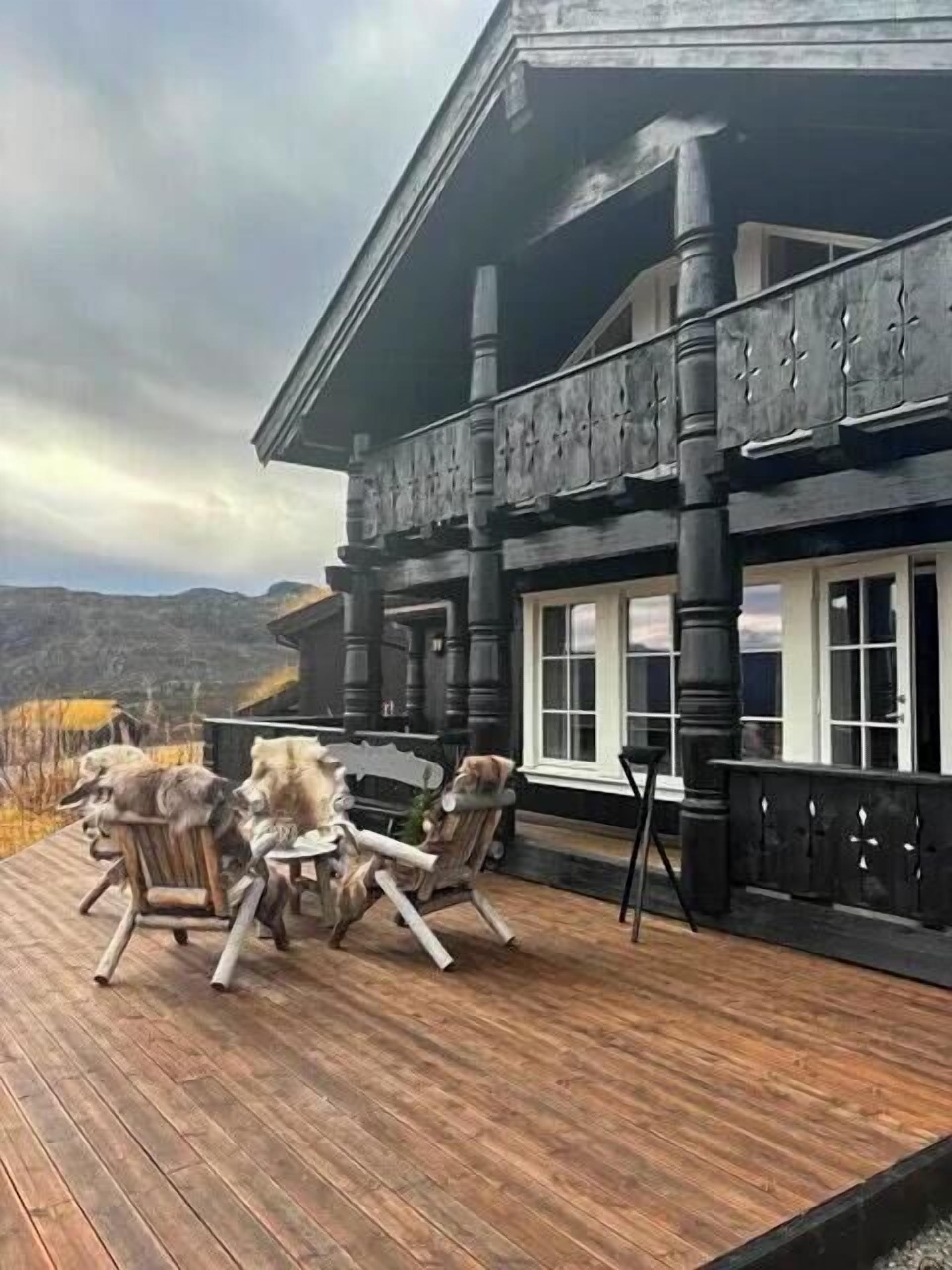 Una terraza de madera con mobiliario exterior, un balcón con barandillas y montañas al fondo.