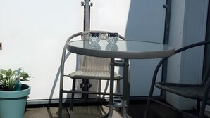 Een klein terras met een ronde glazen tafel en twee stoelen, een plant in pot, en een witte muur op de achtergrond.