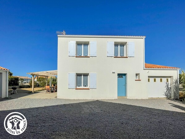 A two-story building with a blue door and white shutters, a gravel driveway, and a clear blue sky. - Spacious lodging near beaches - 6 guests (Le Bernard)