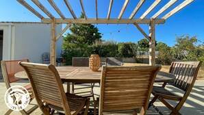 Eine hölzerne Pergola im Freien mit Tisch und Stühlen darunter, umgeben von Grünpflanzen und blauem Himmel.