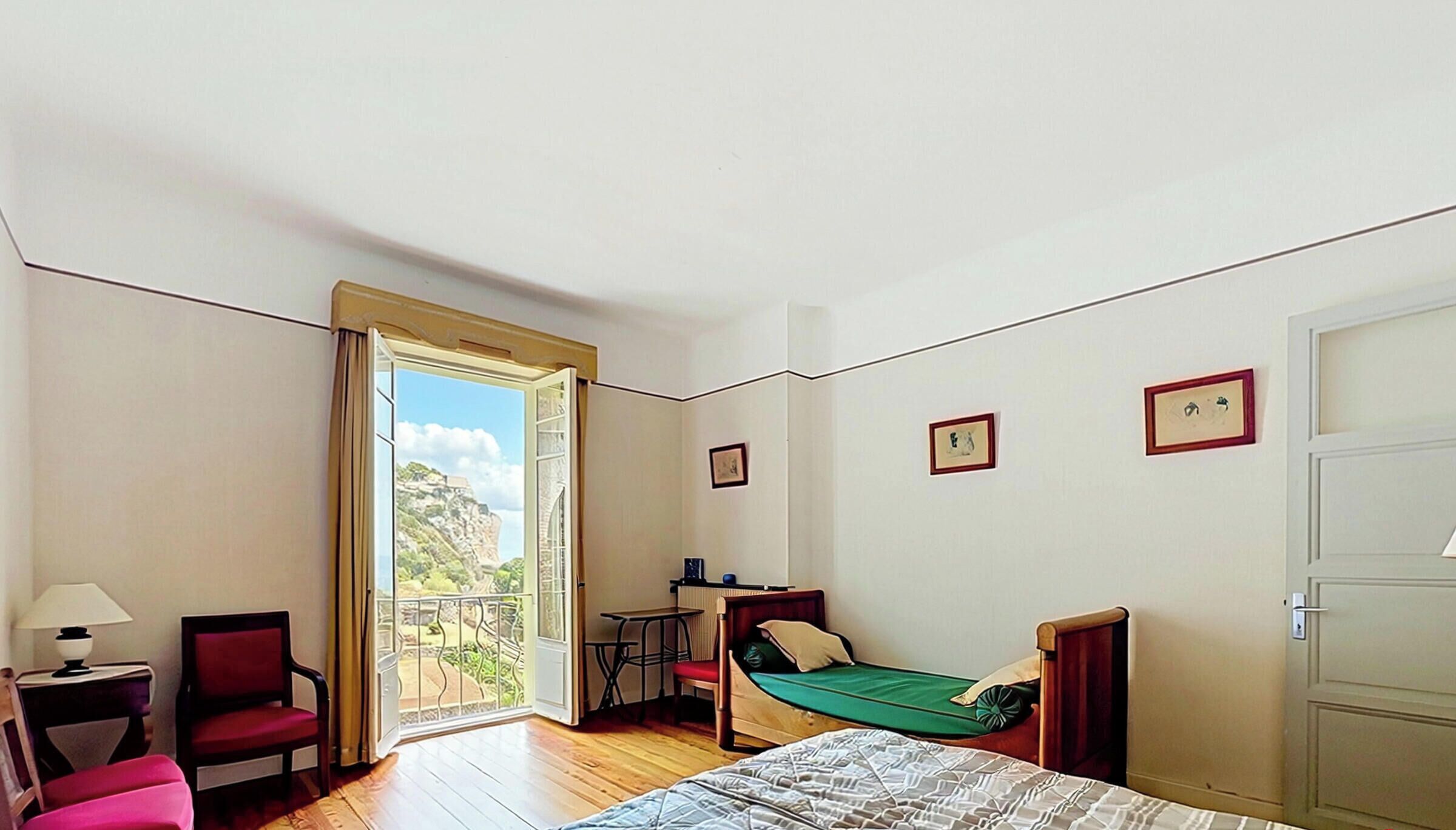A room with a bed, a chair, a table, a lamp, and a balcony with a view.