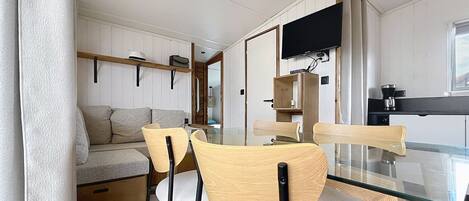 A modern interior with a dining area featuring a glass table and wooden chairs, a sofa, and a television mounted on the wall.