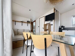 A modern interior with a dining area featuring a glass table and wooden chairs, a sofa, and a television mounted on the wall. - House with parking - 6 guests (Saint-Pair-sur-Mer)