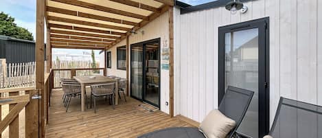 Un patio techado al aire libre con una mesa y sillas de madera, un ventanal de vidrio que da a una habitación y vistas del entorno.
