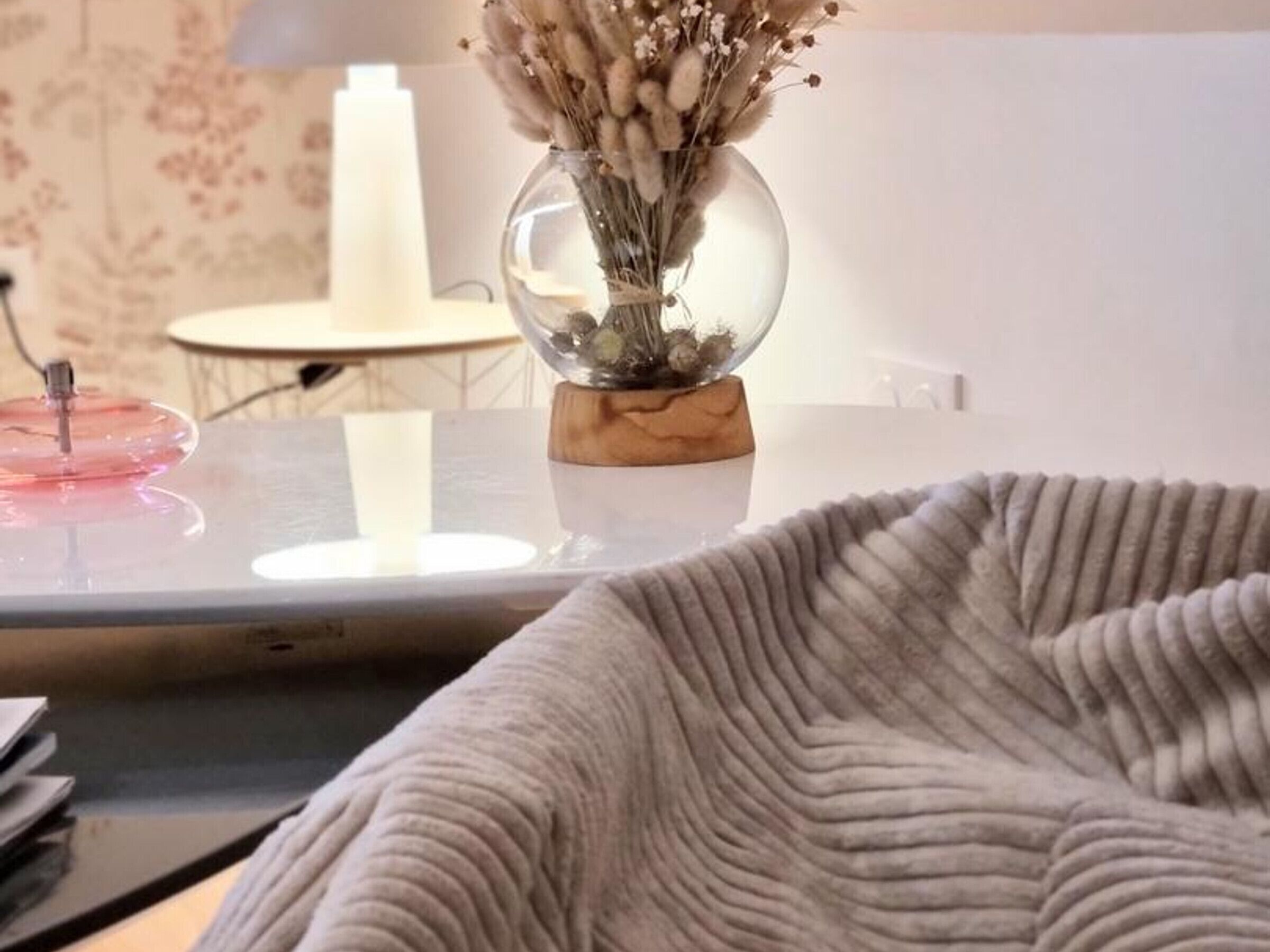 A room with a glass table, a vase with dried flowers, a lamp, and a grey knitted blanket.
