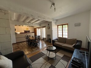 Ein Wohnzimmer mit Sofa, Couchtisch und einem Fernsehschrank. Es gibt einen Essbereich mit Tisch und Stühlen. Der Raum hat einen Fliesenboden und eine Decke mit sichtbaren Balken.