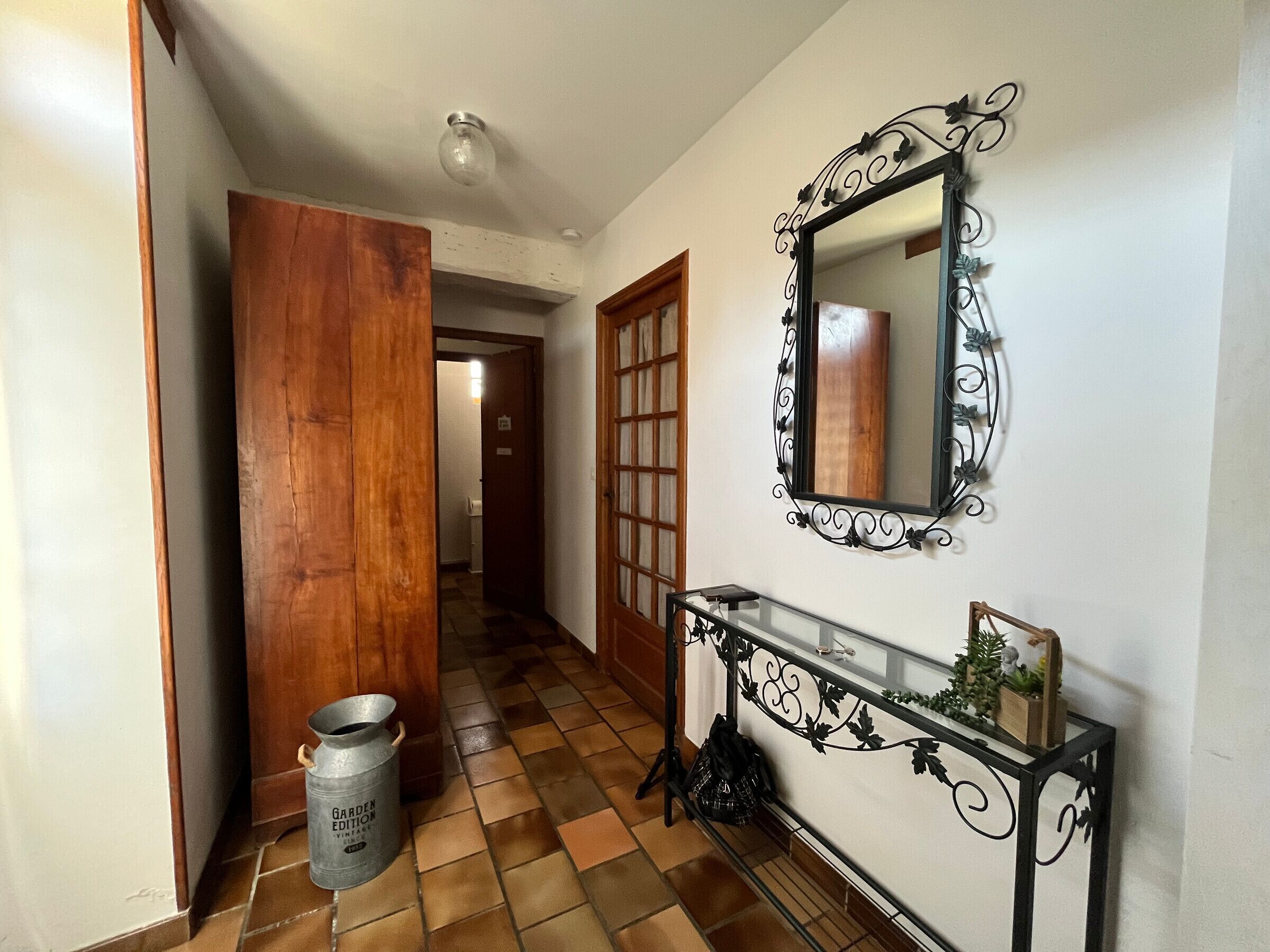 Un corridoio con pavimento piastrellato, una porta in legno, un cestino metallico, un tavolo consolle con uno specchio e uno specchio decorativo fissato alla parete.