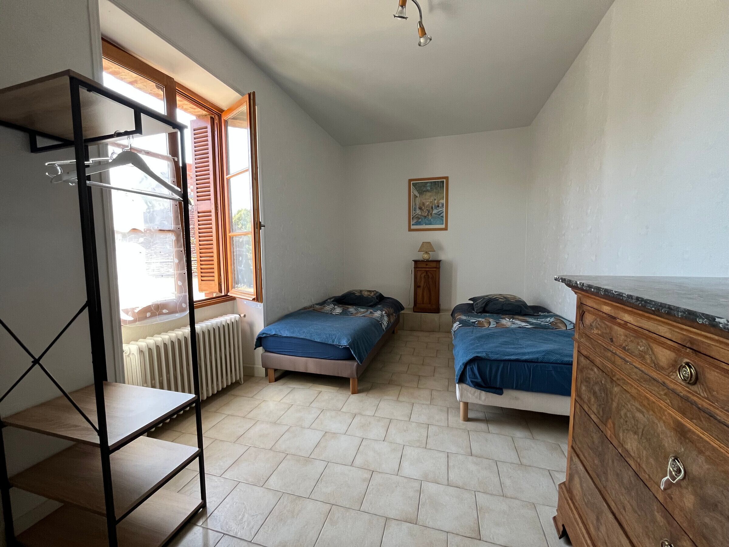 A bedroom with two beds, a radiator, a wooden dresser, and a shelf unit.