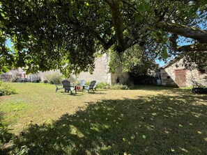Eine grüne Außengelände mit einem Steinhaus, einem Baum, der Schatten spendet, und Gartenmöbeln.