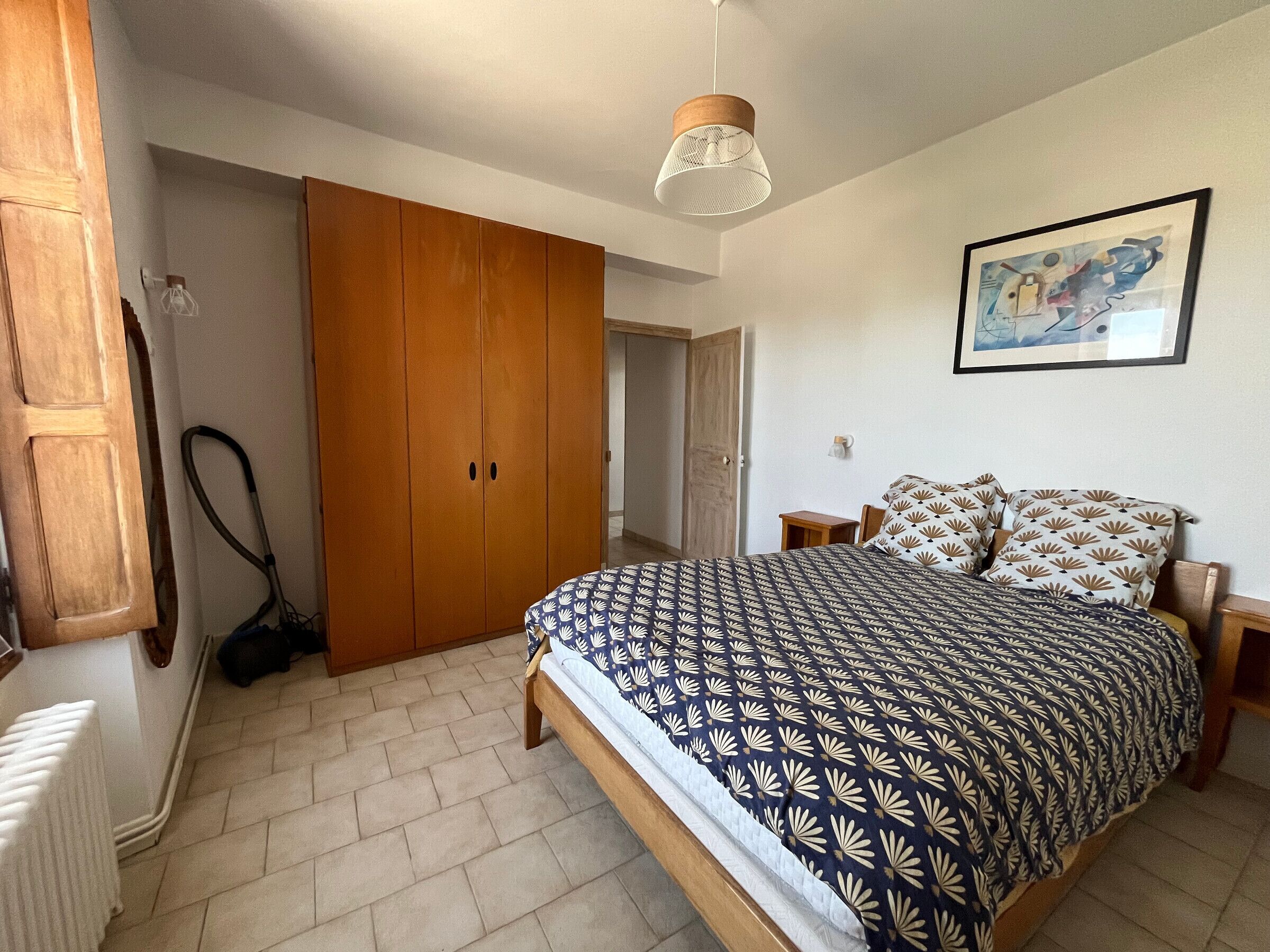Ein Schlafzimmer mit Bett, Holzschrank, Heizkörper, Pendelleuchte und einem gerahmten Bild an der Wand.