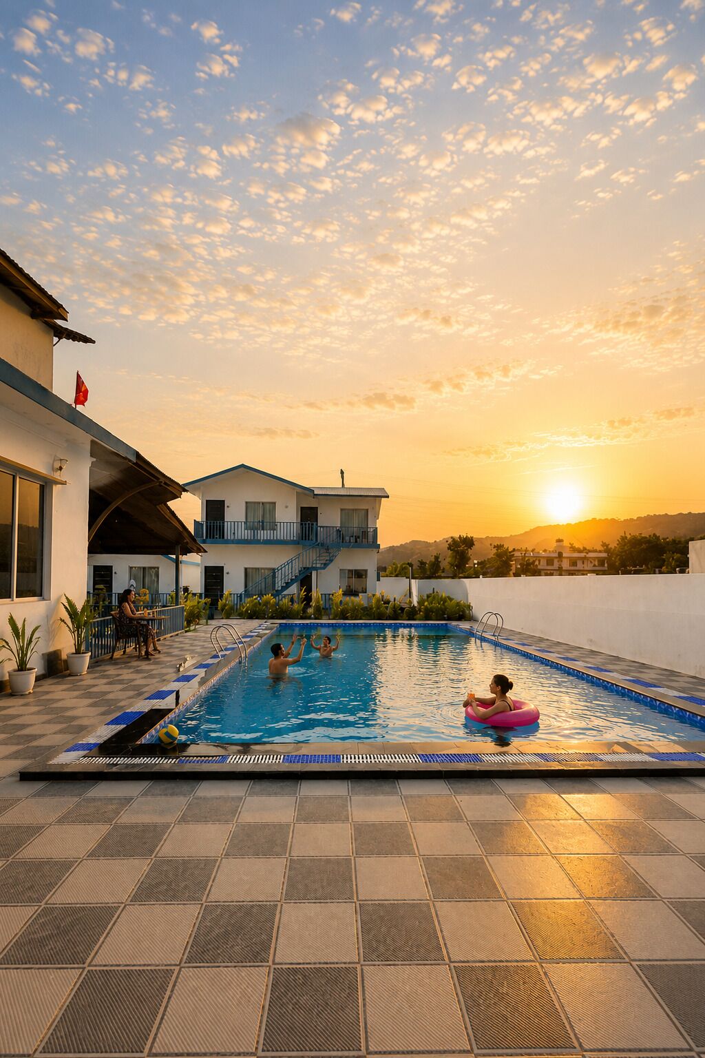 Una piscina con borde de baldosas, un edificio con balcón y una puesta de sol al fondo.