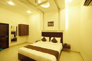 Quarto de hotel com cama, mesas de cabeceira, guarda-roupa, ventoinha no teto e um quadro na parede.