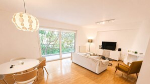A modern living room with a sofa, a dining table with chairs, a TV, and a large window with a view of trees. - Premium 3 Bd Retreat in Neapoli-Lycabettus (Athina)