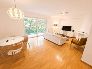 A modern living room with a sofa, a dining table with chairs, a TV, and a large window with a view of trees. - Premium 3 Bd Retreat in Neapoli-Lycabettus (Athina)
