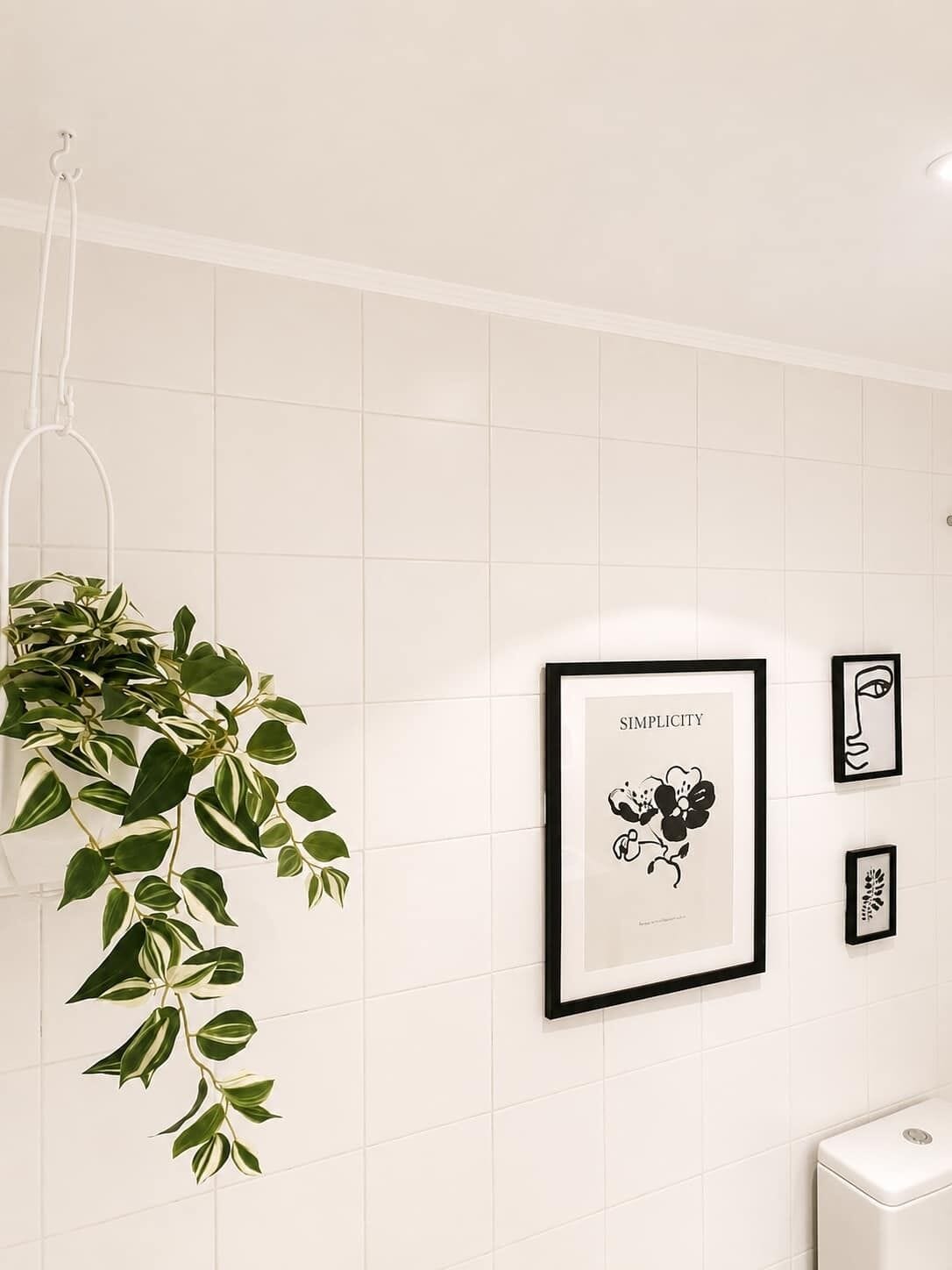 Ein Badezimmer mit weiß gefliesten Wänden, einer hängenden Pflanze und gerahmten Kunstwerken, darunter ein Schwarz-Weiß-Blumendesign und ein gerahmtes Textbild.