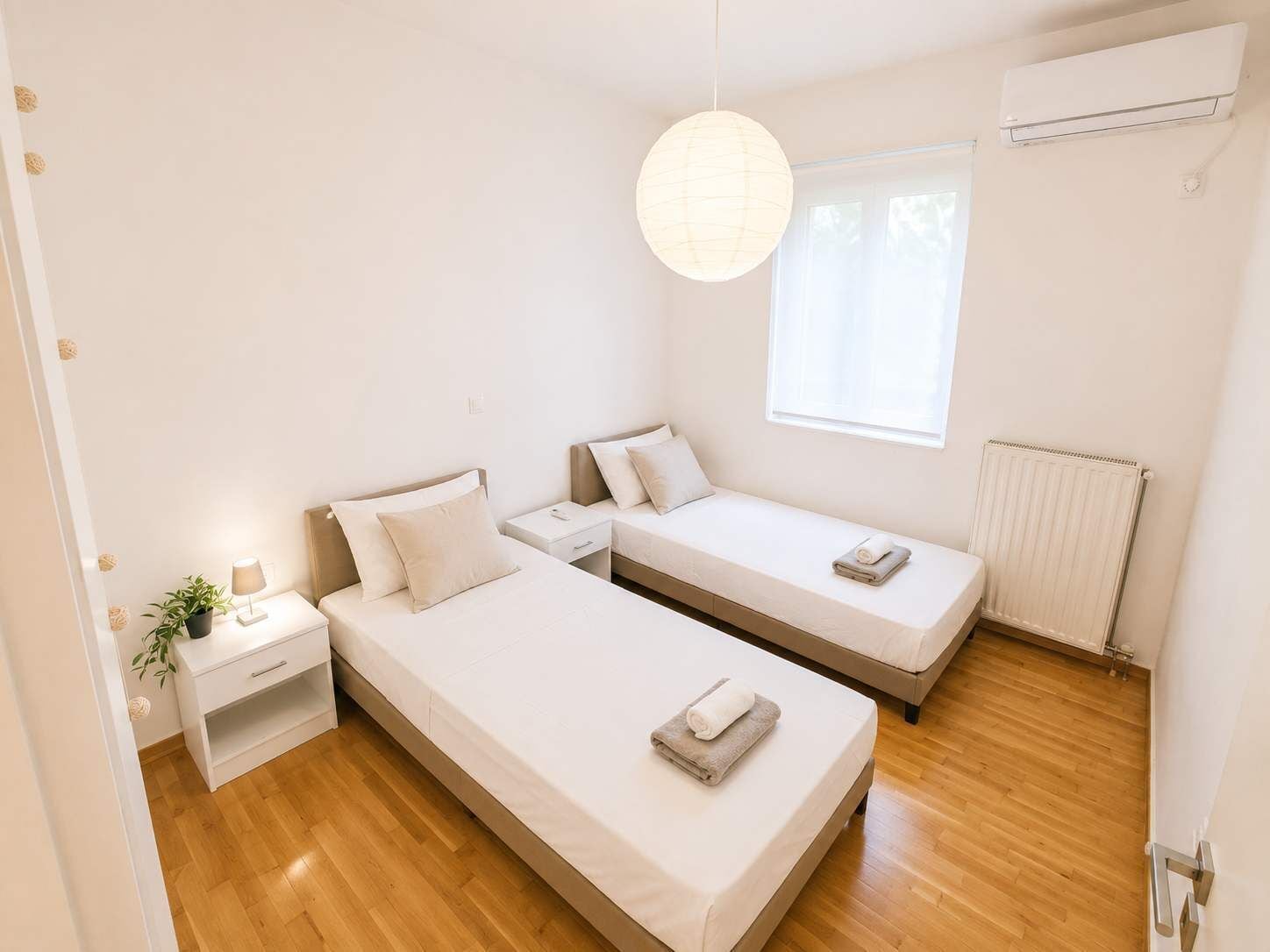 Ein modernes Schlafzimmer mit zwei Einzelbetten, einem Nachttisch mit Lampe, einer Topfpflanze und einem Fenster mit Jalousien.