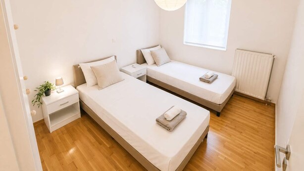A modern bedroom with two single beds, a bedside table with a lamp, a potted plant, and a window with blinds. - Premium 3 Bd Retreat in Neapoli-Lycabettus (Athina)