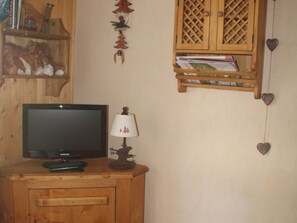 A wooden cabinet with a television and lamp on top, a shelf with books and decorative items, and a wall with a heart-shaped decoration. - Studio near funicular - 4 guests (Bourg-Saint-Maurice)