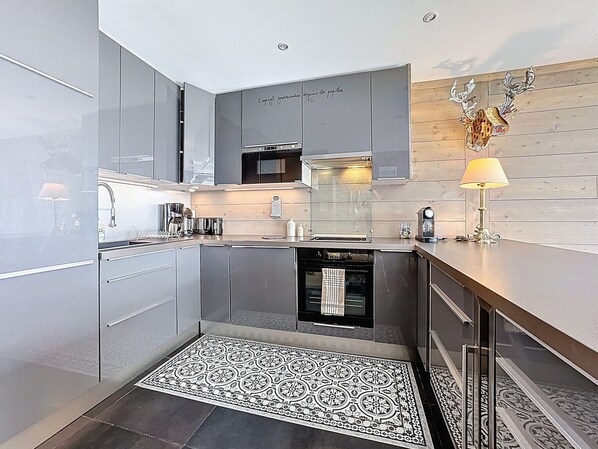 A modern kitchen with grey cabinets, a built-in oven, and a patterned rug. - Spacious apartment, central location - 8 guests (Le Corbier)