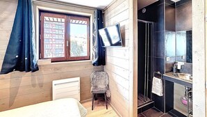Ett sovrum med en säng, en stol, en tv, ett fönster med gardiner och ett badrum med dusch och handfat.