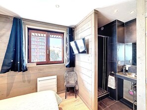 A bedroom with a bed, a chair, a television, a window with curtains, and a bathroom with a shower and sink. - Spacious apartment, central location - 8 guests (Le Corbier)