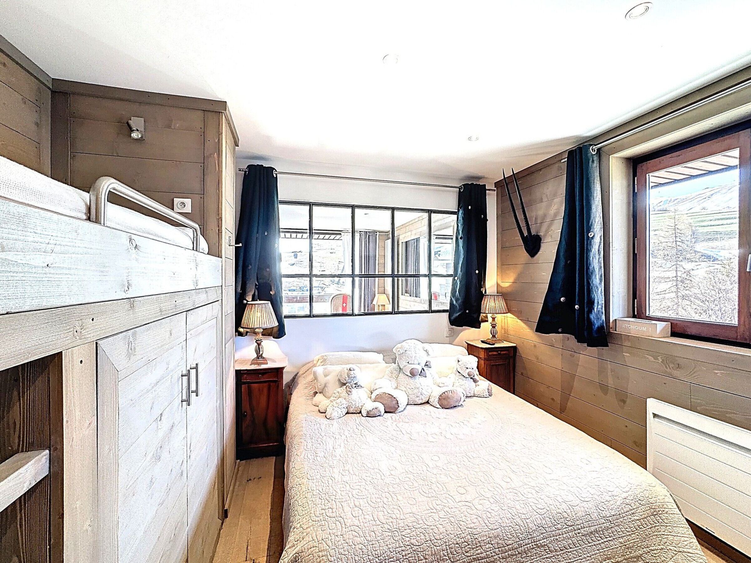 A room with bunk beds, a window with curtains, a bedside table with a lamp, and a view of the outside.