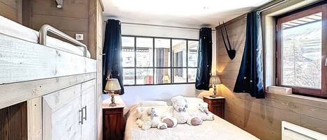 A room with bunk beds, a window with curtains, a bedside table with a lamp, and a view of the outside.