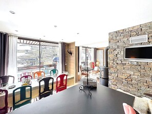 A dining area with a table and chairs, a stone wall, a mounted television, and a window with a view of a mountainous landscape. - Spacious apartment, central location - 8 guests (Le Corbier)