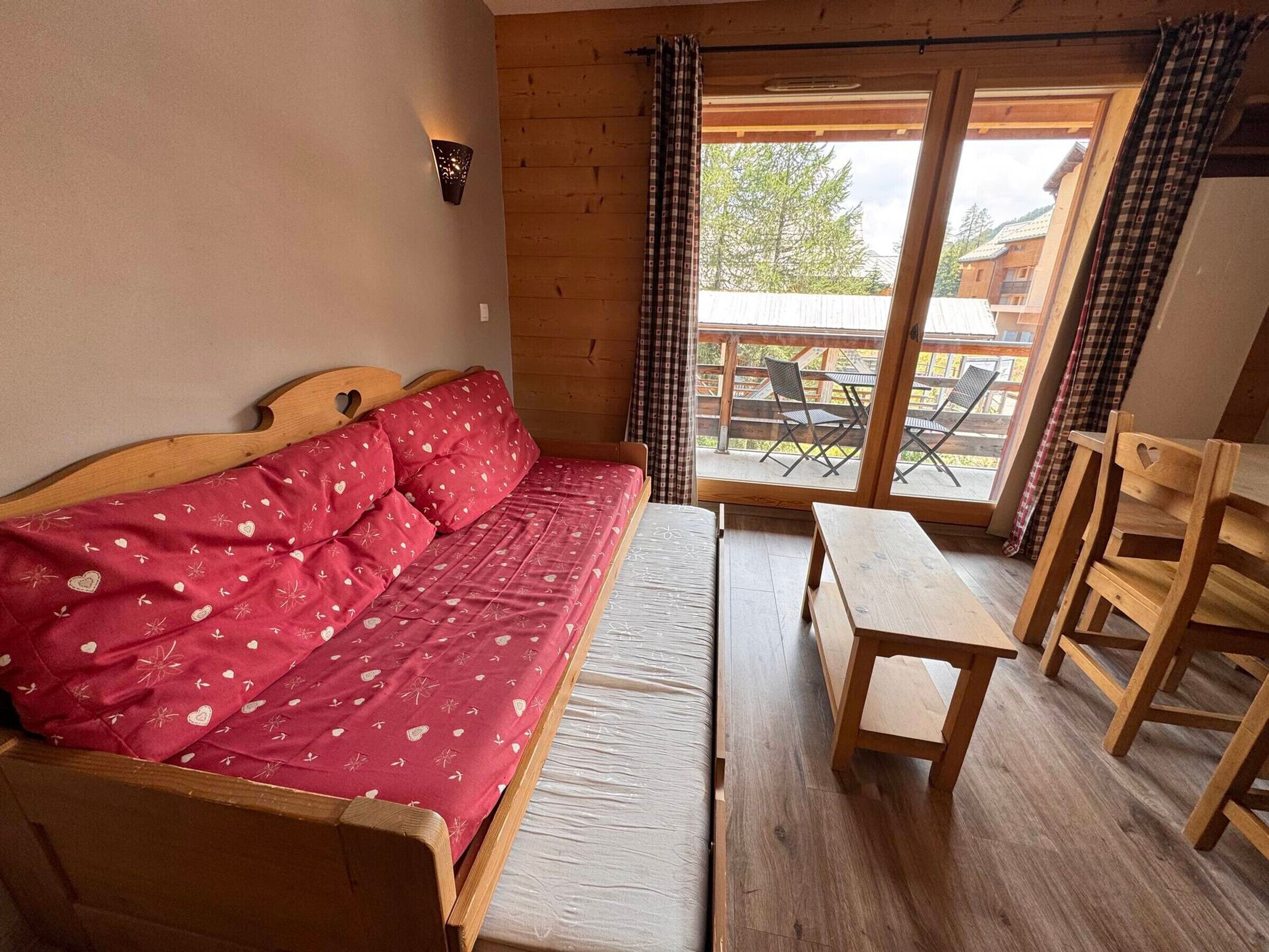 A wooden bed with a red patterned bedspread, a wooden bench, and a table with chairs in a room with wooden walls and a view through a glass door.