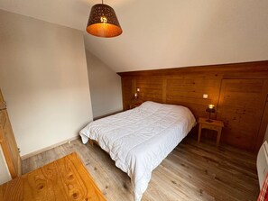 Makuuhuoneessa on puinen sängynpääty, valkoisilla vuodevaatteilla varustettu sänky, yöpöytä lampun kanssa ja puulattia.