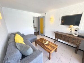 Uma sala de estar com um sofá, uma mesa de centro de madeira, uma televisão na parede e uma pequena mesa com abajur.
