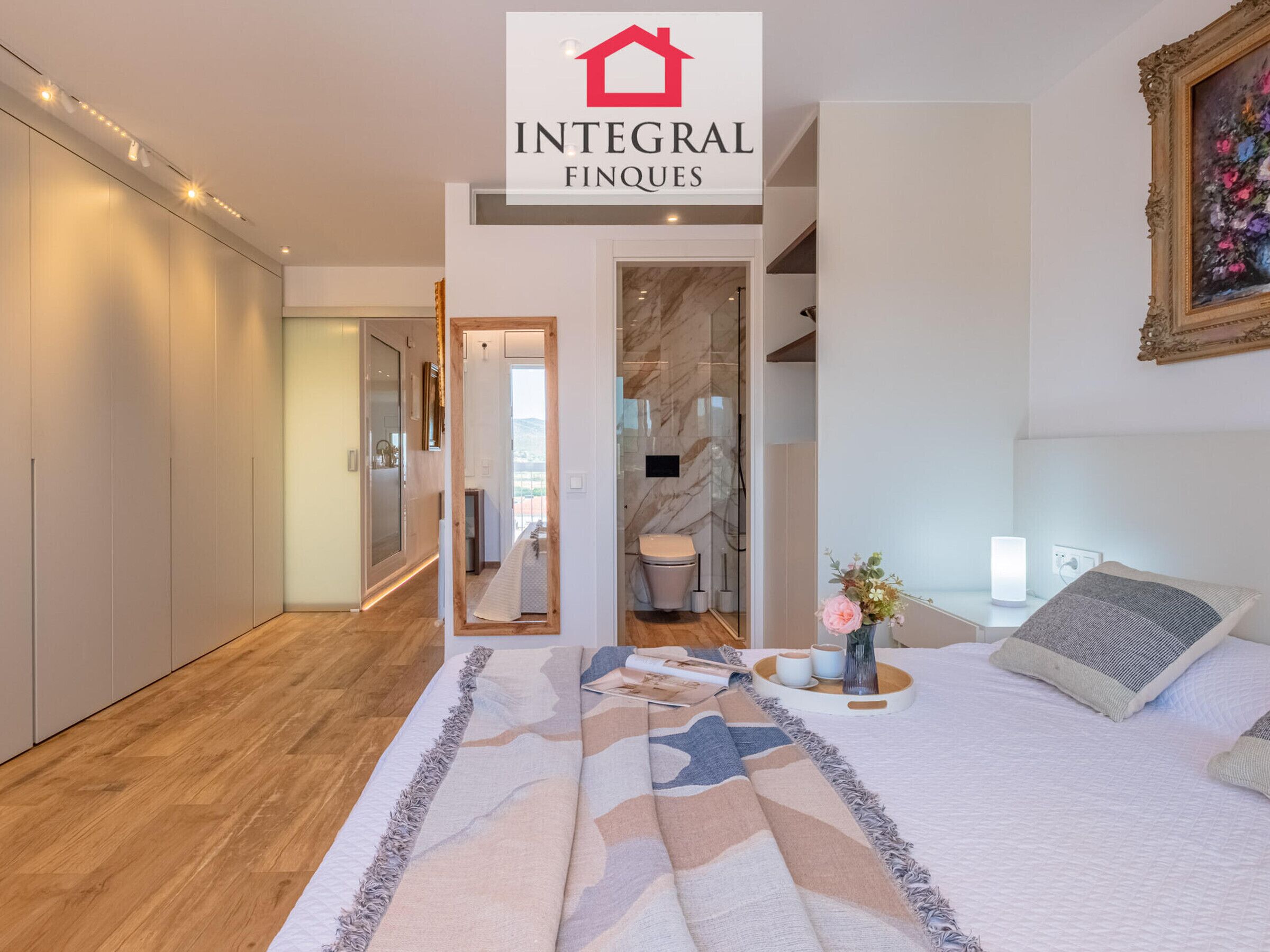 A modern bedroom with a large bed, a bedside table with a lamp, and a mirror. There is a bathroom visible through an open door.