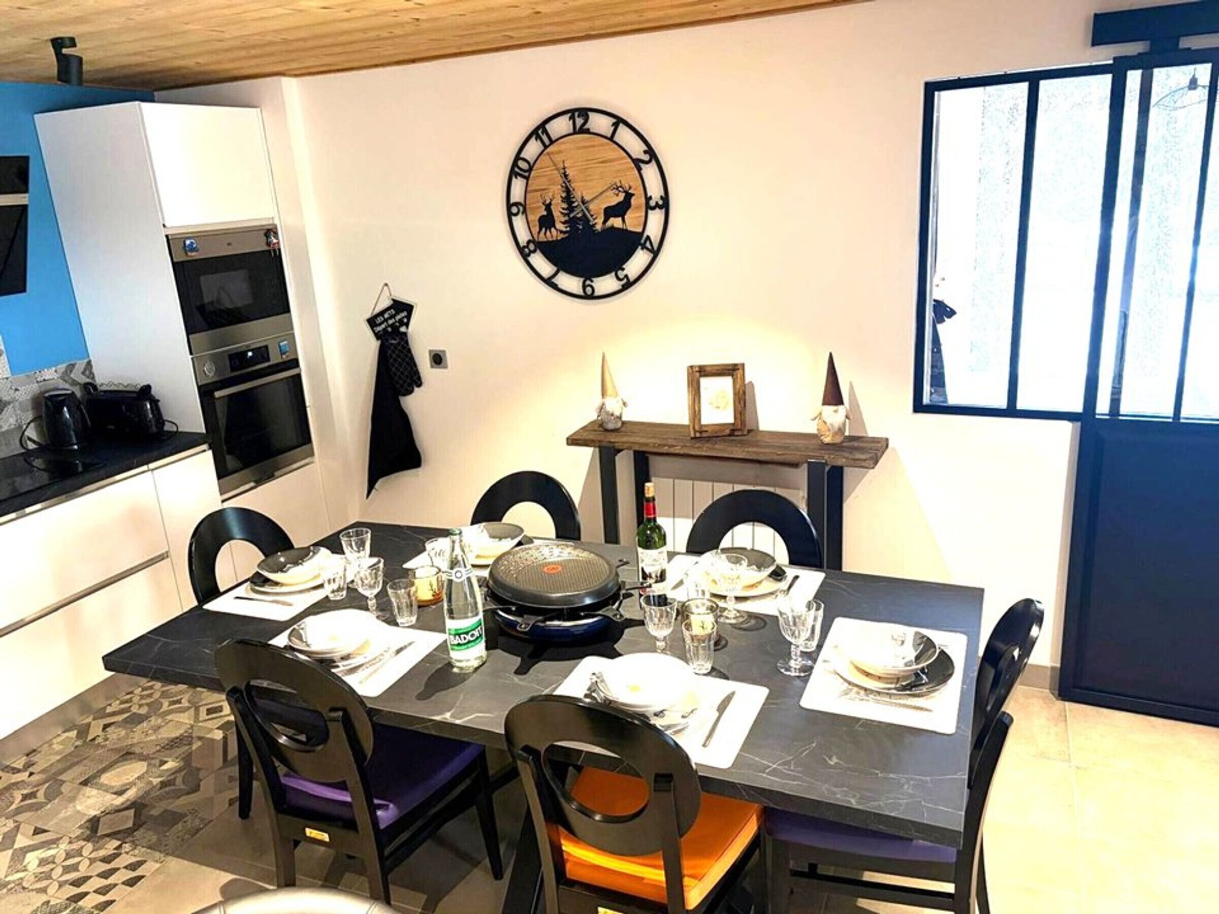 Comedor con una mesa lista para cuatro, sillas, un reloj y una cocina al fondo.
