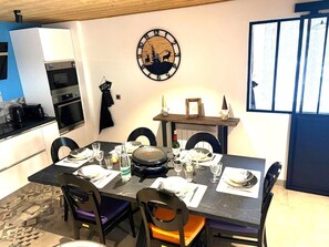 Uma sala de jantar com uma mesa posta para quatro pessoas, cadeiras, um relógio e uma cozinha ao fundo.
