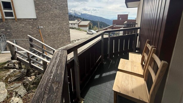 A balcony with a wooden railing, a table, and chairs, overlooking a parking area and buildings with mountains in the background. - Studio cabin with mountain view - 4 guests (Les Adrets-Prapoutel)
