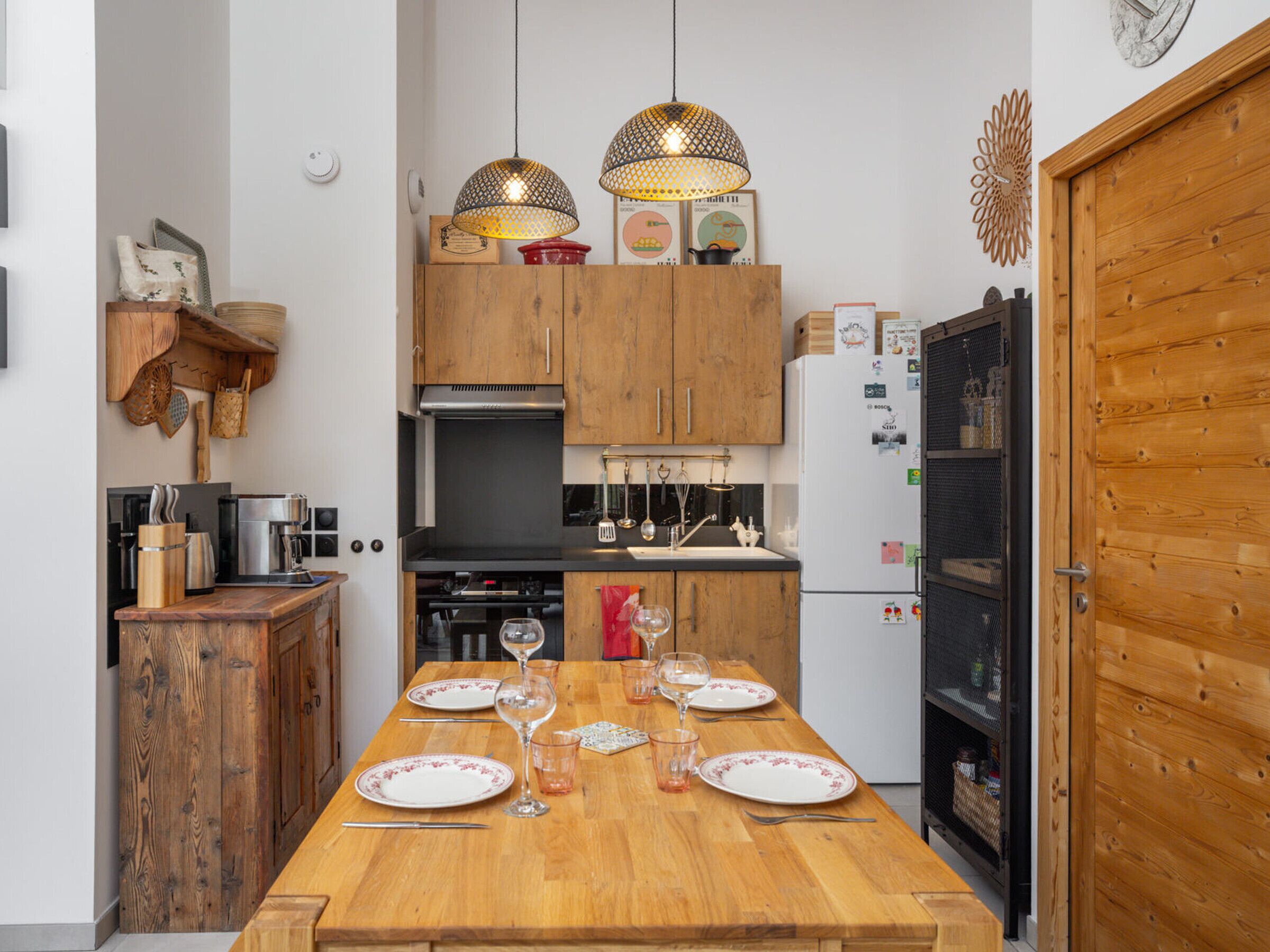 A kitchen with wooden cabinets, a dining table set with plates and glasses, and hanging pendant lights.