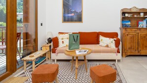 Un salon chaleureux avec un canapé, deux poufs, une petite table, une bibliothèque et une grande fenêtre donnant sur un paysage verdoyant.