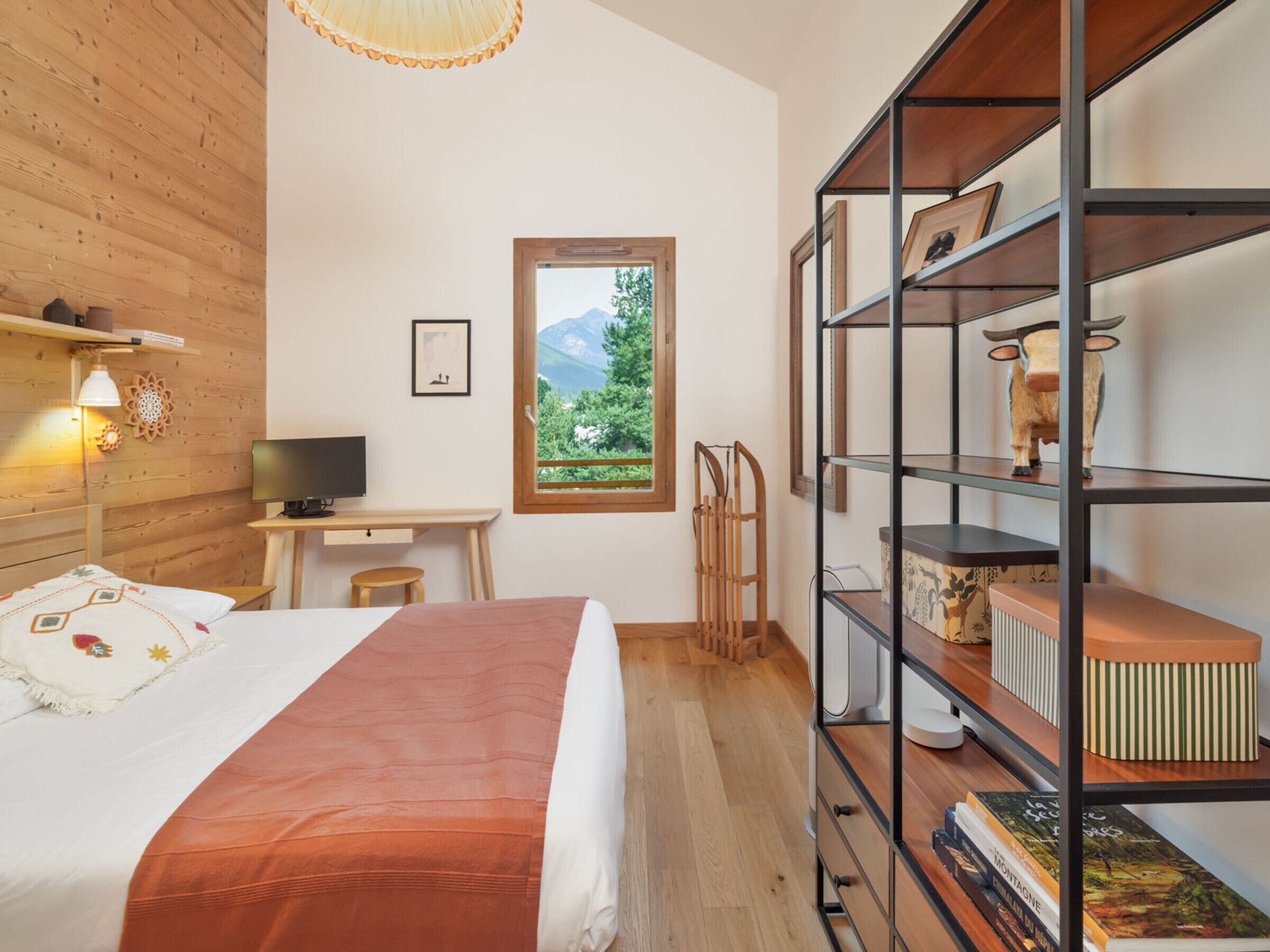 Ein Schlafzimmer mit Bett, Schreibtisch, Stuhl, Bücherregal und einem Fenster mit Bergblick.