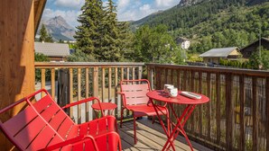 Une terrasse en bois avec des chaises en métal rouge et une petite table, d’où l’on peut admirer un paysage montagneux parsemé de maisons et d’arbres.
