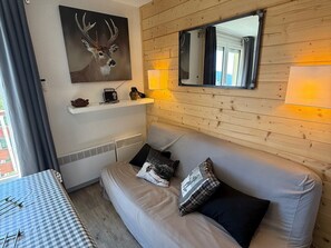 Ett mysigt rum med en väggpanel av trä, en tavla med ett rådjur på väggen, en soffa med kuddar, ett litet bord med en radio och ett fönster med gardiner.
