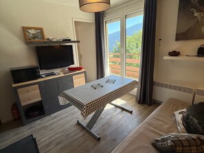 Ein gemütliches Zimmer mit Bett, kleinem Schreibtisch mit Fernseher, einem Fenster mit Vorhängen und Blick ins Grüne.