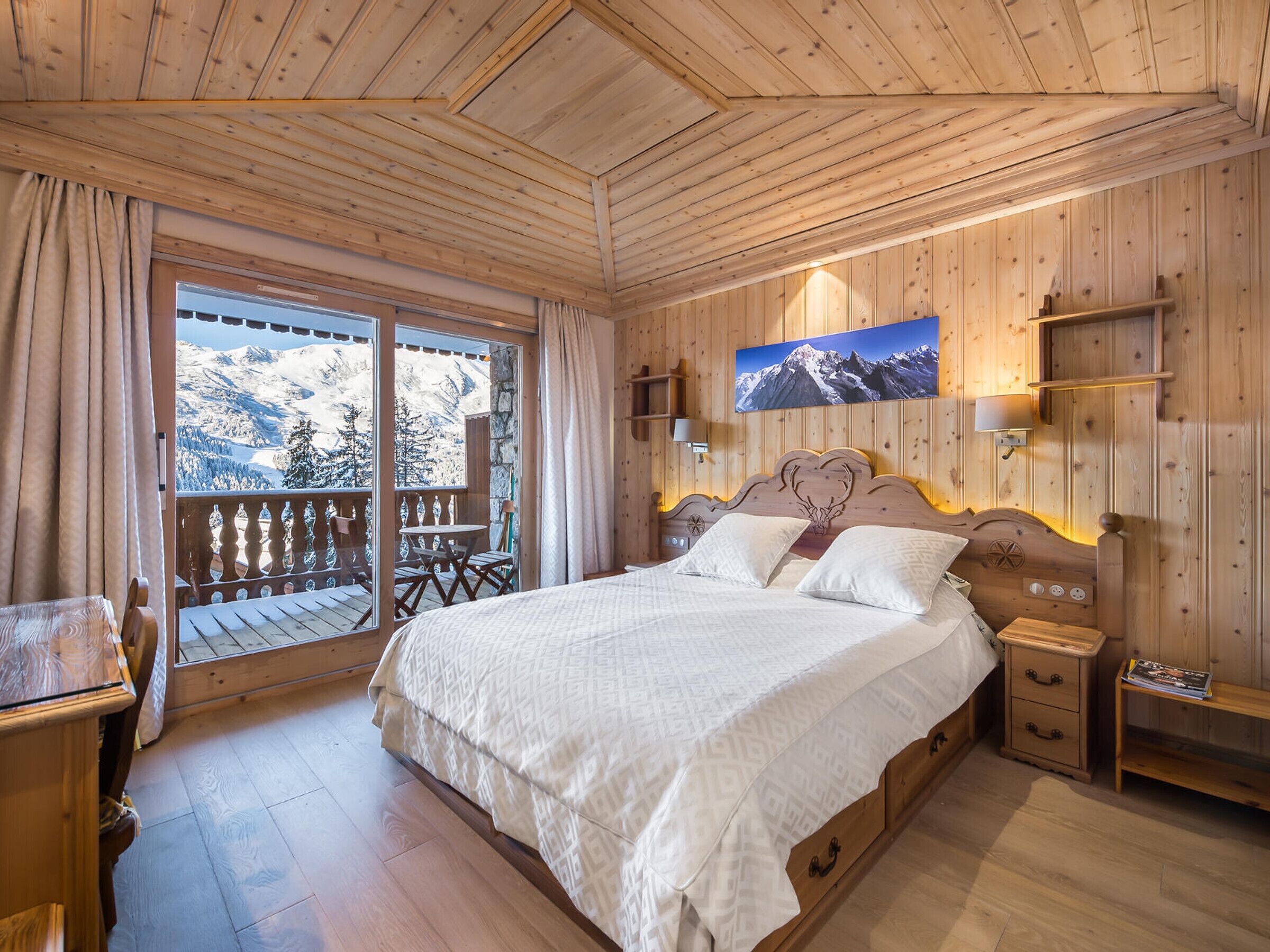 A wooden-paneled bedroom with a bed, bedside tables, a mounted TV, and a balcony with a mountain view.